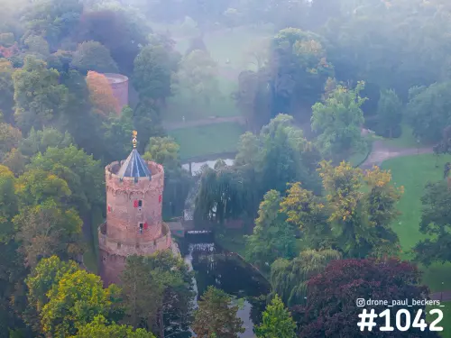 Luchtfoto van een ronde bakstenen toren in het Kronenburgerpark in Nijmegen, met herfstkleuren en mist rond de vijver