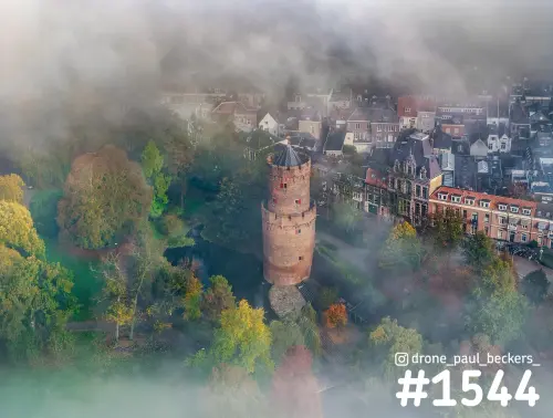 Luchtfoto van een ronde bakstenen toren in het Kronenburgerpark in Nijmegen, met mist tussen herfstbomen en stadshuizen