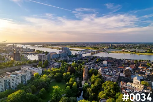 Luchtfoto van Kronenburgerpark in Nijmegen met ronde toren tussen het groen, met de Waal en een boogbrug in laag zonlicht