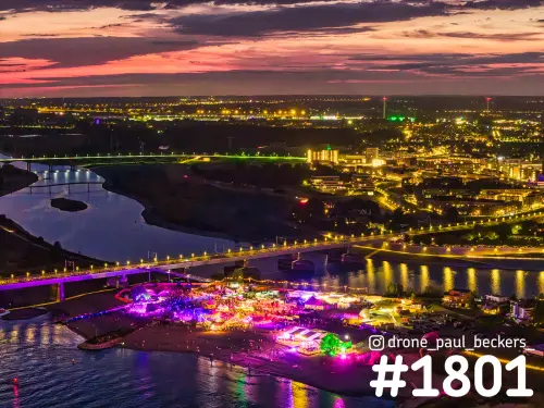 Nijmegen en Lent van boven na zonsondergang, met verlicht festivalterrein op het strand en een brug vol lichtjes