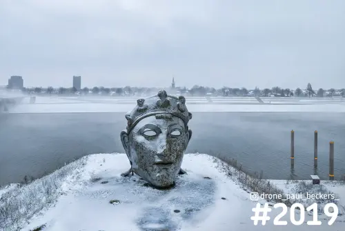 Luchtfoto van een groot gezichtsbeeld met kroon op een besneeuwde oever in Lent, Nijmegen, met mist boven de rivier
