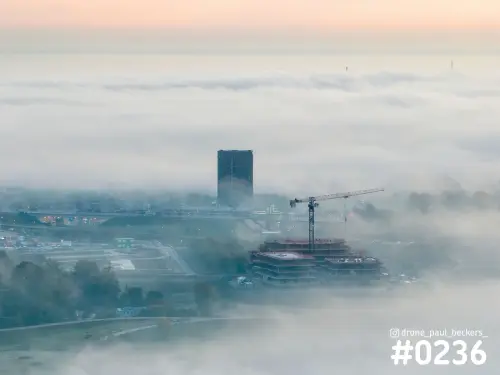 Luchtfoto van Lent in Nijmegen met een donkere toren en een bouwkraan die boven de laaghangende mist uitsteken