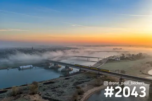 Luchtfoto van een trein op de spoorbrug over de Waal bij Nijmegen-Lent, met mist boven de rivier en een oranje zonsopkomst