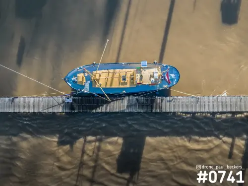 Luchtfoto van een blauw schip aan een houten steiger in de Lindenberghaven in Nijmegen, recht van boven, met lange schaduwen op het water