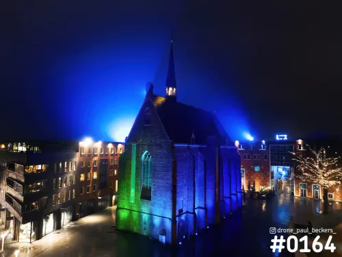 Luchtfoto van de Marienburg kapel in Nijmegen bij nacht, blauw en groen aangelicht in de mist boven het natte plein