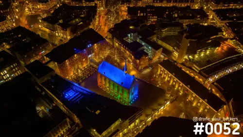 Luchtfoto van de Marienburg kapel in Nijmegen 's avonds, blauw en groen uitgelicht tussen verlichte gevels en kale bomen