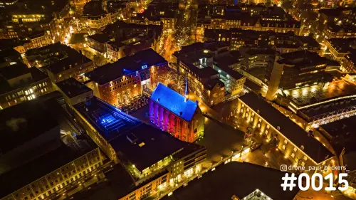 Luchtfoto van de Marienburg kapel in Nijmegen bij avond, met blauw verlichte kap en paarsroze muren tussen stadsgebouwen