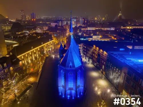 Luchtfoto van de blauw verlichte Marienburg kapel in Nijmegen bij nacht, met lichtsnoeren in de bomen ernaast