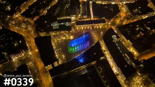 Luchtfoto van de Marienburg kapel in Nijmegen bij nacht, blauw en groen verlicht tussen donkere daken en verlichte straten
