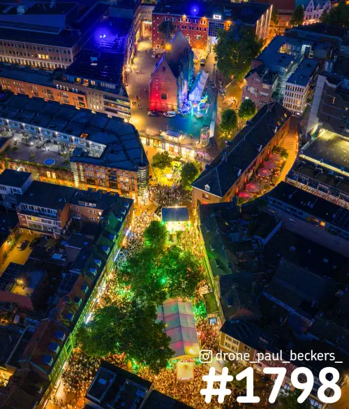 Luchtfoto van de Marienburg kapel in Nijmegen 's avonds, met een menigte onder groen verlichte bomen