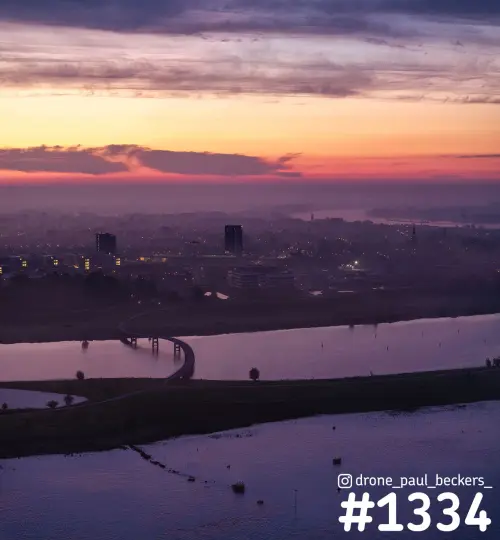 Luchtfoto van de Waal en de nevengeul bij Nijmegen-Lent in de avondgloed, met een gebogen brug en mist boven de stad