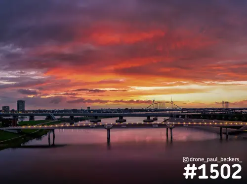 Luchtfoto van een verlichte brug over de nevengeul bij Nijmegen-Lent onder een rood-oranje wolkenlucht in de schemering