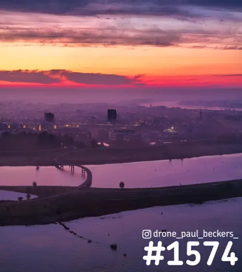 Luchtfoto van de nevengeul bij Nijmegen-Lent in de ochtendschemering, met een gebogen brug en mist boven de stad