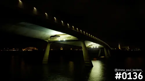 Luchtfoto van een betonnen brug bij de Nevengeul in Nijmegen 's avonds, met verlichte rand en Stevenskerk in de verte