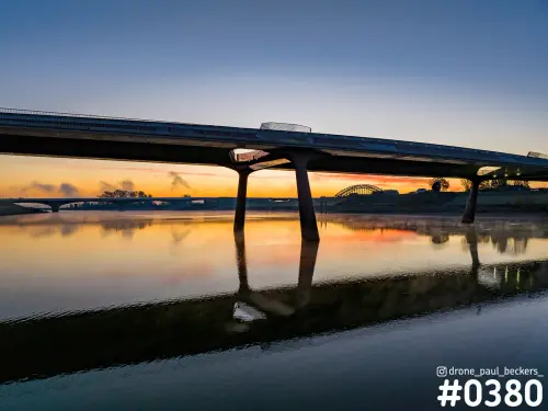 Betonnen brug over de Nevengeul in Nijmegen in oranje schemerlicht, met spiegeling in het water en boogbrug op de horizon
