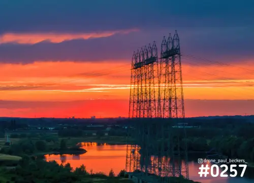 Hoogspanningsmasten aan de Nevengeul bij Nijmegen tijdens een rood-oranje zonsondergang, met reflectie in het water