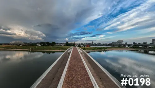 Luchtfoto van de voetgangersbrug over de Nevengeul in Nijmegen, met dreigende wolkenlucht en weerspiegeling in het water
