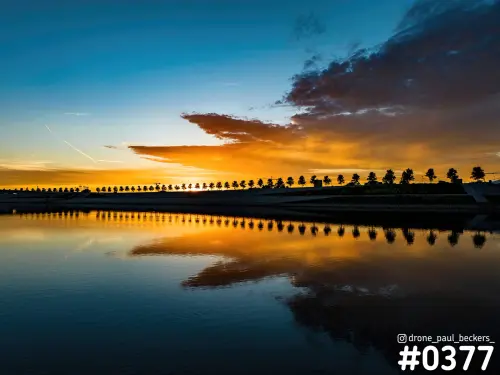Rij bomen als silhouet langs de Nevengeul bij zonsondergang, met oranje wolken en de weerspiegeling in het stille water