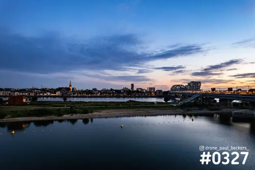Luchtfoto van de nevengeul en de Waal in Nijmegen bij schemering, met stadslichten, gele boeien en een brug rechts