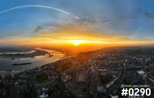 Luchtfoto van het centrum van Nijmegen bij zonsopkomst, met de Stevenskerk, de Waal en de Waalbrug onder een oranje gloed