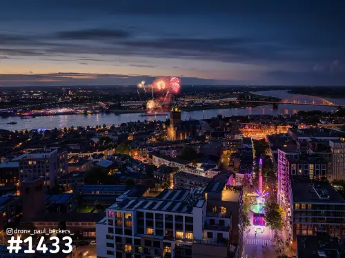 Luchtfoto van Nijmegen in de avondschemering met vuurwerk boven de Waal, de Stevenskerk en een verlichte kermisattractie