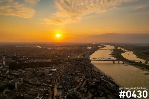 Luchtfoto van het centrum van Nijmegen en de Waal bij zonsondergang, met een oranje lucht en een brug over de rivier