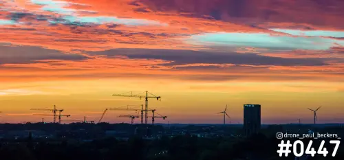 Panorama van Nijmegen-Lent in de schemering met bouwkranen, een hoteltoren en windturbines tegen een oranje wolkenlucht