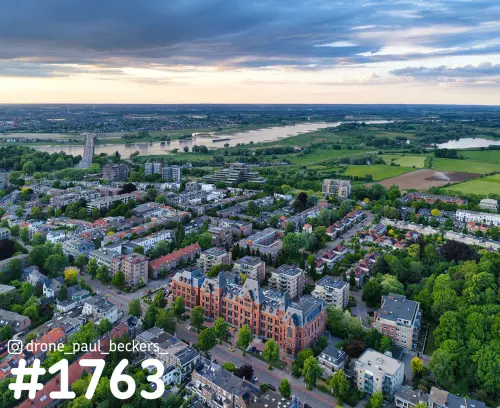 Luchtfoto van Nijmegen-Oost met een rood bakstenen gebouw, de Waal, de Waalbrug en de Ooijpolder bij zonsondergang