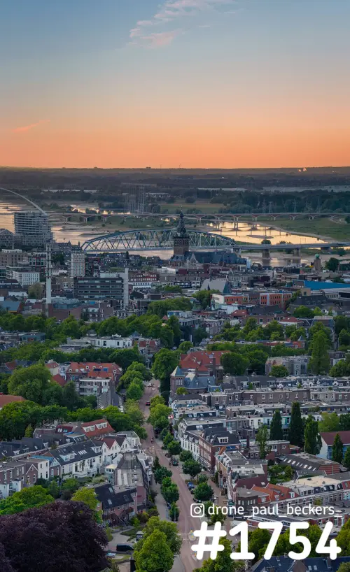Luchtfoto van Nijmegen met Stevenskerk en de Waal, woonwijk met bomen op de voorgrond en oranje gloed aan de horizon