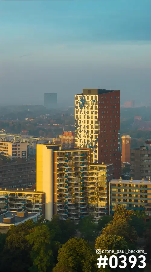 Luchtfoto van de Nimbus-toren in Nijmegen in warm ochtendlicht, met gele gevel, rode baksteenzijde en nevel boven de stad
