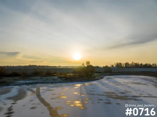 Luchtfoto van een bevroren plas in de Ooijpolder met laagstaande zon, gouden weerspiegelingen op het ijs en kale bomen