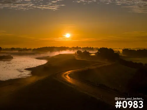 Luchtfoto van de Ooijpolder bij zonsopkomst, met een slingerende dijkweg en mist boven het water onder een oranje lucht