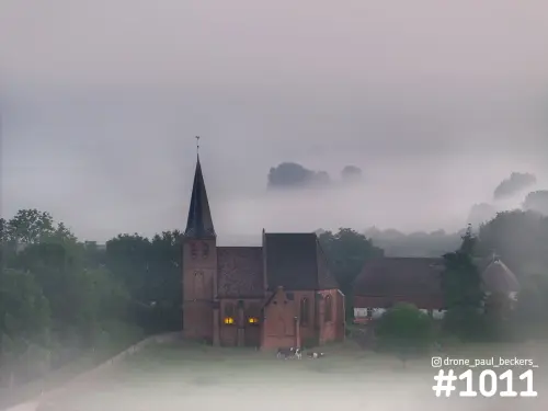 Luchtfoto van een bakstenen kerk met spitse toren in de Ooijpolder, met mist, verlichte ramen en koeien in de wei