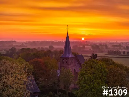 Luchtfoto van een kerk met spitse toren in de Ooijpolder bij zonsopkomst, met oranje lucht en mist boven de velden