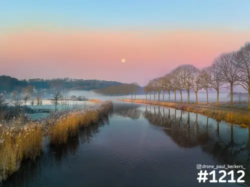 Luchtfoto van de Ooijpolder bij Nijmegen met mist, een rij kale bomen langs het water en een volle maan in roze ochtendlucht