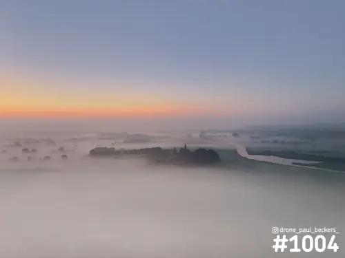 Luchtfoto van de Ooijpolder bij Nijmegen in de ochtendmist, met oranje gloed aan de horizon en een kerktoren boven de mist