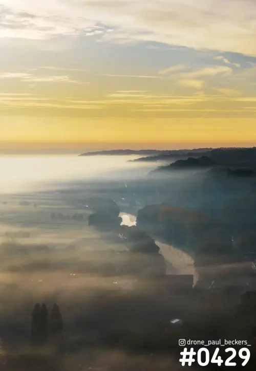Luchtfoto van de Ooijpolder in de mist bij zonsopkomst, met een rivierbocht en bosrand onder een gele lucht