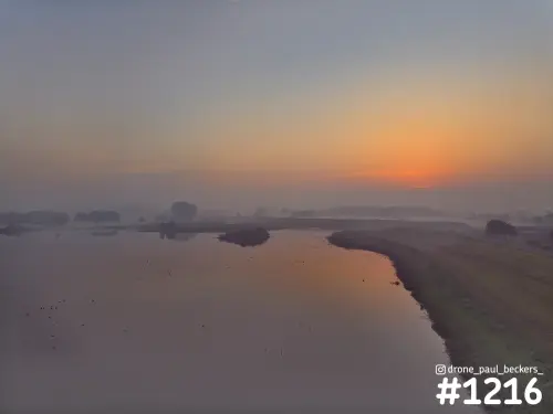 Luchtfoto van de Ooijpolder bij Nijmegen: oranje gloed aan de horizon, mist boven het water en een grazige oever rechts