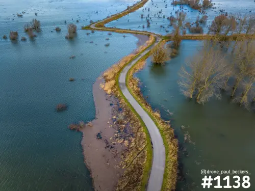 Luchtfoto van een smalle asfaltweg die als een dijkje door het overstroomde landschap van de Ooijpolder slingert, met kale bomen in het water