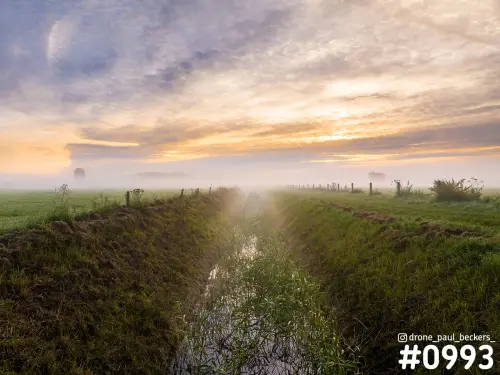 Luchtfoto van een sloot in de Ooijpolder bij zonsopkomst, met mist boven weilanden en een oranje wolkenlucht