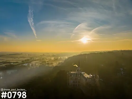 Luchtfoto van de Ooijpolder bij zonsopkomst met mist over de velden en een bakstenen gebouw met torenspits op een beboste heuvel