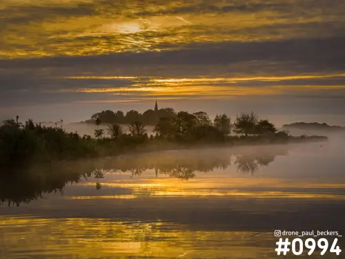 Luchtfoto van de Ooijpolder bij zonsopkomst, met gouden wolken en een kerktorentje achter de mist boven het water