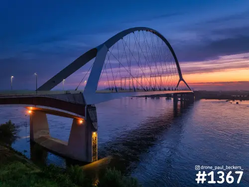 Luchtfoto van de Oversteek over de Waal bij Nijmegen-Lent in de schemering, met oranje horizon en verlichte brugpijler