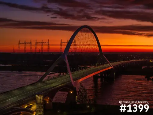 Luchtfoto van de Oversteek in Nijmegen bij zonsondergang, met de boog van de brug en hoogspanningsmasten tegen een oranjerode lucht
