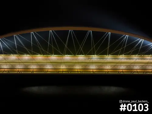 Luchtfoto van de Oversteek in Nijmegen bij nacht, met de verlichte brugboog, kabels en lichtsporen van verkeer over het wegdek