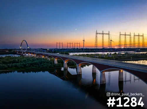 Luchtfoto van De Oversteek in Nijmegen bij zonsondergang, met de brug gespiegeld in de nevengeul en hoogspanningsmasten