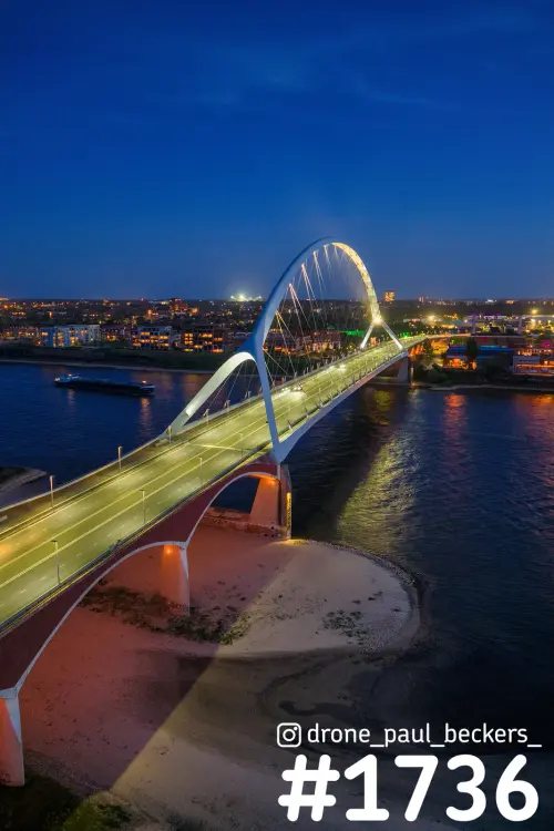 Luchtfoto van De Oversteek in Nijmegen bij avondschemering, met verlichte boogbrug en een zandbank in de Waal