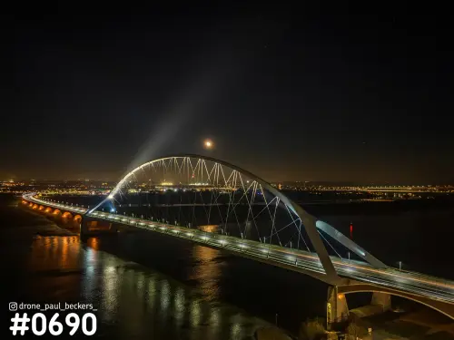 Luchtfoto van de Oversteek over de Waal bij Nijmegen in de nacht, met verlichte boogbrug en maan boven de rivier