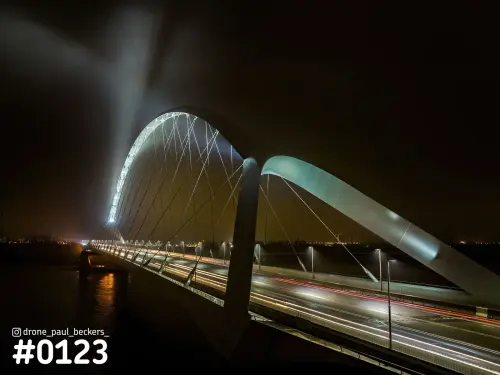 Luchtfoto van de boog van De Oversteek over de Waal bij Nijmegen in de nacht, met lichtstrepen van het verkeer en mist