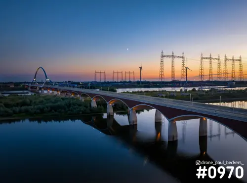 Luchtfoto van De Oversteek in Nijmegen na zonsondergang, met de boogbrug over de Waal, hoogspanningsmasten en maan